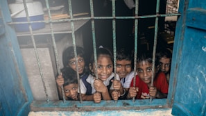 A group of young children smiling through a window with metal bars. The children appear cheerful and lively, looking directly at the viewer. The blue window frame contrasts with the children's colorful clothing.
