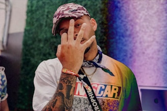 A person wearing a printed t-shirt and a camouflage cap makes a gesture with their hand in front of their face. Tattoos are visible on the forearm, and a bandana is tied around the neck. The background shows a colorful, textured wall with shades of green and purple.