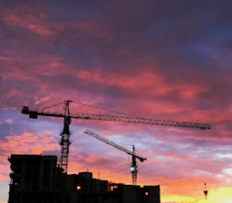 A powerful purple and saffron hydraulic crane lifting heavy steel beams at a busy Mumbai construction site during sunset.