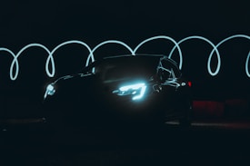 A dark setting with a black car illuminated by its headlights, creating a striking visual. Behind the car, wavy light trails glow, suggesting motion or long exposure photography.