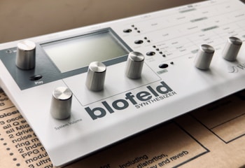 A compact digital synthesizer with multiple knobs and a small display. The brand name 'Blofeld' and the word 'SYNTHESIZER' are prominently visible. It is placed on a cardboard surface.