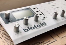 A compact digital synthesizer with multiple knobs and a small display. The brand name 'Blofeld' and the word 'SYNTHESIZER' are prominently visible. It is placed on a cardboard surface.