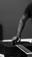 A person is tuning or adjusting the strings of an acoustic guitar. The focus is on the hand and part of the forearm, while the background is plain and blurred, adding an artistic feel. The image is in black and white, enhancing the contrasts and shadows.
