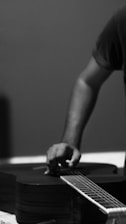 A person is tuning or adjusting the strings of an acoustic guitar. The focus is on the hand and part of the forearm, while the background is plain and blurred, adding an artistic feel. The image is in black and white, enhancing the contrasts and shadows.