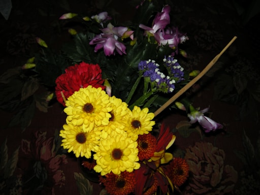 A vibrant arrangement of assorted flowers featuring yellow daisies, red carnations, purple orchids, and small violet flowers, set against dark green leaves. The flowers are placed on a dark floral-patterned surface.