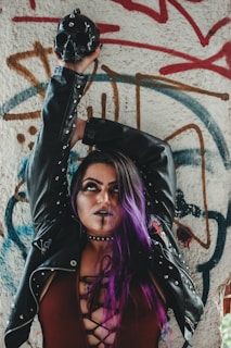 A person with purple hair and dark makeup wearing a black leather jacket stands in front of a graffiti-covered wall. They are holding a black skull with spikes above their head. The jacket has metal studs, and the background features various colorful graffiti designs in red, brown, and blue.