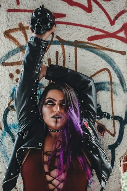 A person with purple hair and dark makeup wearing a black leather jacket stands in front of a graffiti-covered wall. They are holding a black skull with spikes above their head. The jacket has metal studs, and the background features various colorful graffiti designs in red, brown, and blue.