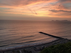 A breathtaking view of the ocean at sunset with soft waves gently reaching the shore. The sky is painted with vibrant hues of orange, pink, and purple, while a dark rocky pier stretches into the sea. A line of vehicles is visible along the coastal road, bordered by a row of palm trees.