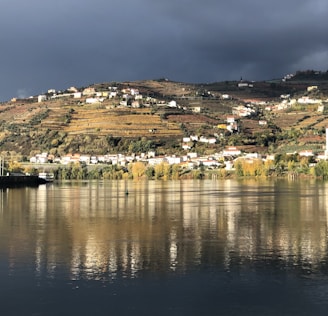 A picturesque landscape featuring a small town with predominantly white buildings nestled on a hillside. The hill is lined with terraced vineyards, indicating a region known for wine production. The foreground shows a calm body of water reflecting the hillside and cloudy sky above, with patches of sunlight illuminating parts of the landscape.