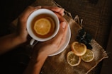 A pair of hands gently holds a white cup filled with tea and lemon slices. The cup rests on a rustic, woven fabric placed on a wooden surface. Beside the cup are additional lemon slices and some dark green leaves.