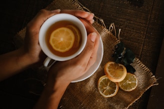A pair of hands gently holds a white cup filled with tea and lemon slices. The cup rests on a rustic, woven fabric placed on a wooden surface. Beside the cup are additional lemon slices and some dark green leaves.