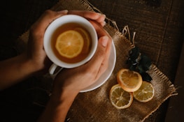 Close-up of hands holding a cup of herbal tea beside an open journal on a white linen cloth.