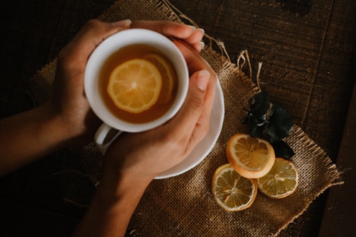 A pair of hands gently holds a white cup filled with tea and lemon slices. The cup rests on a rustic, woven fabric placed on a wooden surface. Beside the cup are additional lemon slices and some dark green leaves.