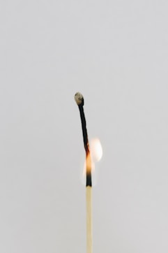 A single used matchstick is shown with a charred, blackened tip, emitting a small flame. The background is a minimalist, plain light gray, creating a stark contrast with the darkened matchstick.