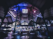A spacious indoor market with ornate metalwork and high ceilings. The central area features a small food stall adorned with various decorations. Several ceiling lights illuminate the area, casting a cool, dim ambiance. Tables are scattered around the space, and people are seen interacting at the food stall.