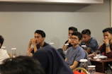 Several young individuals are seated at tables in a conference or classroom setting. They appear attentive and engaged, some with thoughtful expressions as they listen or contemplate the session in progress. The tables are equipped with glasses and documents, possibly indicating a seminar or educational event.