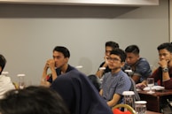 Several young individuals are seated at tables in a conference or classroom setting. They appear attentive and engaged, some with thoughtful expressions as they listen or contemplate the session in progress. The tables are equipped with glasses and documents, possibly indicating a seminar or educational event.