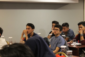 Several young individuals are seated at tables in a conference or classroom setting. They appear attentive and engaged, some with thoughtful expressions as they listen or contemplate the session in progress. The tables are equipped with glasses and documents, possibly indicating a seminar or educational event.