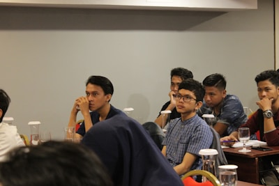 Several young individuals are seated at tables in a conference or classroom setting. They appear attentive and engaged, some with thoughtful expressions as they listen or contemplate the session in progress. The tables are equipped with glasses and documents, possibly indicating a seminar or educational event.