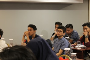 Several young individuals are seated at tables in a conference or classroom setting. They appear attentive and engaged, some with thoughtful expressions as they listen or contemplate the session in progress. The tables are equipped with glasses and documents, possibly indicating a seminar or educational event.