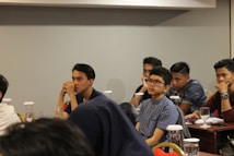 Several young individuals are seated at tables in a conference or classroom setting. They appear attentive and engaged, some with thoughtful expressions as they listen or contemplate the session in progress. The tables are equipped with glasses and documents, possibly indicating a seminar or educational event.