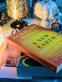 A cozy, moody atmosphere with a stack of books on a table beside decorative items. Prominently featured is 'A New Earth' by Eckhart Tolle, with a yellow cover. The setting is illuminated by a lamp with intricate patterns and soft, ambient lighting.