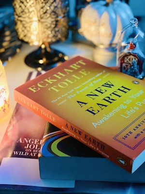 A cozy, moody atmosphere with a stack of books on a table beside decorative items. Prominently featured is 'A New Earth' by Eckhart Tolle, with a yellow cover. The setting is illuminated by a lamp with intricate patterns and soft, ambient lighting.