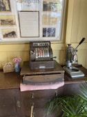 An antique cash register is placed on a wooden cabinet alongside a vintage coffee machine. Behind them, a framed historical display featuring sepia-toned photographs and text is mounted on a cream-colored wall. There is also a small vase with pink flowers and a box labeled 'My Family Tree' containing sticks with labels.