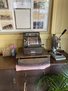 An antique cash register is placed on a wooden cabinet alongside a vintage coffee machine. Behind them, a framed historical display featuring sepia-toned photographs and text is mounted on a cream-colored wall. There is also a small vase with pink flowers and a box labeled 'My Family Tree' containing sticks with labels.