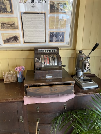 An antique cash register is placed on a wooden cabinet alongside a vintage coffee machine. Behind them, a framed historical display featuring sepia-toned photographs and text is mounted on a cream-colored wall. There is also a small vase with pink flowers and a box labeled 'My Family Tree' containing sticks with labels.