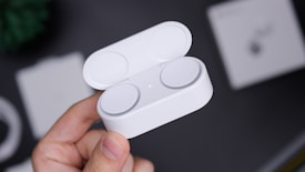 A hand holding a small, white, oval-shaped case that is open to reveal two earbuds or circular electronic devices inside. The background is blurred, with various objects that appear indistinct.