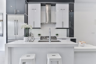 A modern kitchen with sleek white and gray cabinetry, featuring a large central island with a built-in sink. Stainless steel appliances, including a refrigerator and range hood, complement the design. Two white stools are positioned at the island, and a vase with white flowers adds a decorative touch. A bowl of fruit is positioned on the countertop, adding a hint of color.