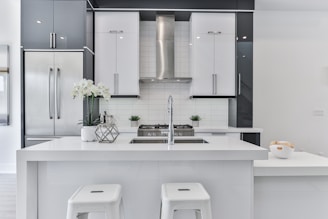 A modern kitchen with sleek white and gray cabinetry, featuring a large central island with a built-in sink. Stainless steel appliances, including a refrigerator and range hood, complement the design. Two white stools are positioned at the island, and a vase with white flowers adds a decorative touch. A bowl of fruit is positioned on the countertop, adding a hint of color.