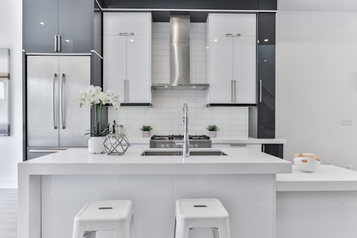 A modern kitchen with sleek white and gray cabinetry, featuring a large central island with a built-in sink. Stainless steel appliances, including a refrigerator and range hood, complement the design. Two white stools are positioned at the island, and a vase with white flowers adds a decorative touch. A bowl of fruit is positioned on the countertop, adding a hint of color.