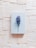 A light blue hardcover book is centrally placed on a textured background composed of overlapping sheets of off-white paper. A single purple and white flower with a green stem is laid across the book, adding a touch of natural beauty and contrast to the scene.