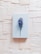 A light blue hardcover book is centrally placed on a textured background composed of overlapping sheets of off-white paper. A single purple and white flower with a green stem is laid across the book, adding a touch of natural beauty and contrast to the scene.