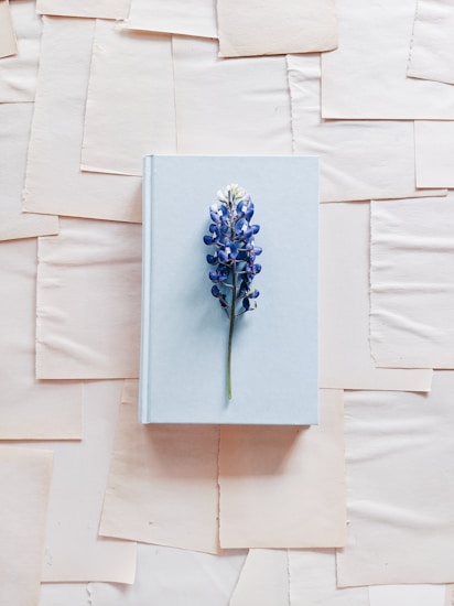 A light blue hardcover book is centrally placed on a textured background composed of overlapping sheets of off-white paper. A single purple and white flower with a green stem is laid across the book, adding a touch of natural beauty and contrast to the scene.