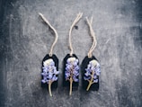 Three dark labels with strings are arranged side by side on a textured surface. Each label features a small bouquet of purple and white flowers attached to it.