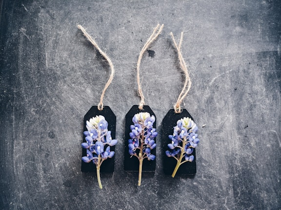 Three dark labels with strings are arranged side by side on a textured surface. Each label features a small bouquet of purple and white flowers attached to it.