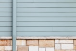 A wall with horizontal siding in a pale blue-gray color is topped with a series of beige and light brown stone bricks. The clean, straight lines of the paneling contrast with the natural texture and patterns of the stonework.