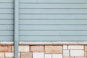 A wall with horizontal siding in a pale blue-gray color is topped with a series of beige and light brown stone bricks. The clean, straight lines of the paneling contrast with the natural texture and patterns of the stonework.
