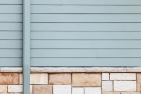 A wall with horizontal siding in a pale blue-gray color is topped with a series of beige and light brown stone bricks. The clean, straight lines of the paneling contrast with the natural texture and patterns of the stonework.