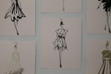The image contains a series of fashion sketches displayed on a wall. Each sketch features abstract, stylistic drawings of women in various fashionable dresses. The artwork is monochromatic and uses a minimalistic design approach.