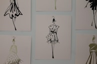 The image contains a series of fashion sketches displayed on a wall. Each sketch features abstract, stylistic drawings of women in various fashionable dresses. The artwork is monochromatic and uses a minimalistic design approach.
