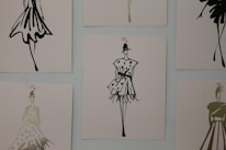 The image contains a series of fashion sketches displayed on a wall. Each sketch features abstract, stylistic drawings of women in various fashionable dresses. The artwork is monochromatic and uses a minimalistic design approach.