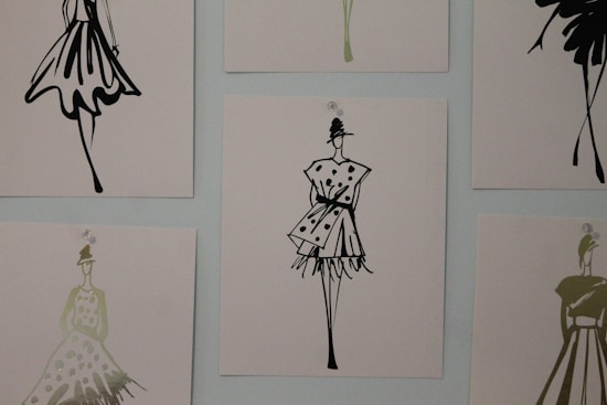 The image contains a series of fashion sketches displayed on a wall. Each sketch features abstract, stylistic drawings of women in various fashionable dresses. The artwork is monochromatic and uses a minimalistic design approach.
