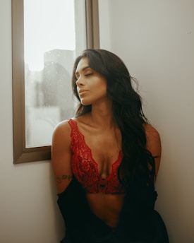 A woman with long dark hair and closed eyes is wearing a red lace top and sitting by a window. The soft lighting creates a serene and elegant atmosphere.