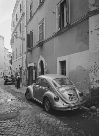 A narrow cobblestone street in Rome with a small car parked near ancient ruins.