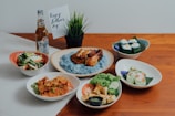 A table set with various dishes including grilled chicken on a bed of blue rice, spring rolls, a bowl of vegetables, and prawns with garnish. There is a bottle of beer and a small card with 'happy father's day' written on it, along with a small potted plant.