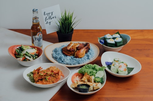 A table set with various dishes including grilled chicken on a bed of blue rice, spring rolls, a bowl of vegetables, and prawns with garnish. There is a bottle of beer and a small card with 'happy father's day' written on it, along with a small potted plant.